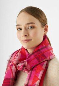Jeune femme à la peau claire et aux yeux bleus portant un foulard à motifs rouges et roses et un haut beige, souriant légèrement sur un fond blanc.