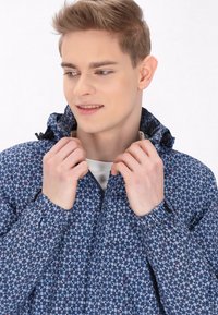 Jachetă albastră cu model, cu guler înalt, dotată cu fermoar și manșete elastice. Materialul pare ușor, cu un design texturat.