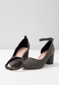 Paire de chaussures à talon bloc en daim gris foncé avec bouts pointus et brides de cheville amovibles, présentées sur une surface blanche.
