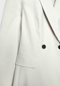 Light grey blazer with a smooth texture, featuring a double-breasted design, black buttons, and a single flap pocket on the side.