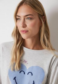 Lichtgrijze t-shirt met een grote blauwe hartafbeelding en een lachend gezicht. Zachte stof, casual ontwerp, ronde halslijn en korte mouwen.