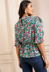 Ziedu blūze zaļā krāsā ar rozā un zilām ziedkopām, ar savilktu jostasvietu, īsām piedurknēm un atslēgas formas atveri aizmugurē, saskaņota ar tumši zilām džinsām.