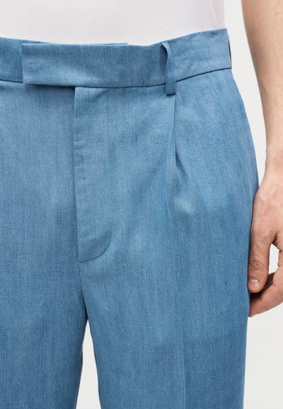 Homme portant un pantalon bleu clair sur mesure avec une ceinture, des passants et une poche latérale, assorti à une chemise blanche.