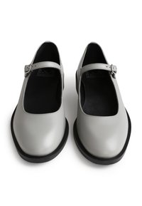 Graue Leder-Mary-Jane-Schuhe mit runder Zehenpartie, flacher schwarzer Sohle und verstellbarem Riemen. Glatte Textur, einfaches Design und minimale Details.
