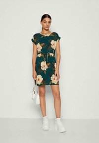 Φλοράλ φόρεμα με μωβ φόντο, κοντά μανίκια και μέση με κορδόνι. Συνδυάζεται με λευκά sneakers και μια μικρή λευκή τσάντα.