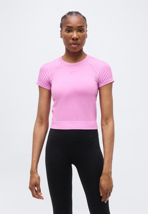 Naine patsidega juustega, kes kannab vormis roosat lühikeste varrukatega sportlikku tuba ja kõrge vöökohaga musti retuuse, seisab näoga ettepoole, neutraalne ilme.