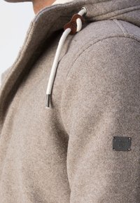 Veste à capuche en laine beige avec cordon blanc et écusson de marque en métal sur le haut du bras, vue de l'épaule et de côté.