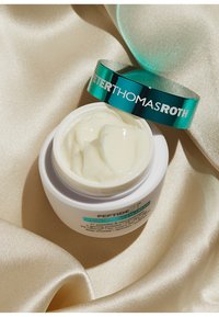 Un pot blanc de crème avec un couvercle teal affichant "PETER THOMAS ROTH." La crème est épaisse et brillante, posée sur un tissu beige soyeux.