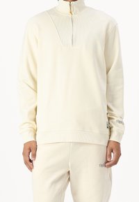 Maglione color crema con collo a mezza zip, trama a coste sul colletto, polsini elastici e marchio discreto sull'orlo.