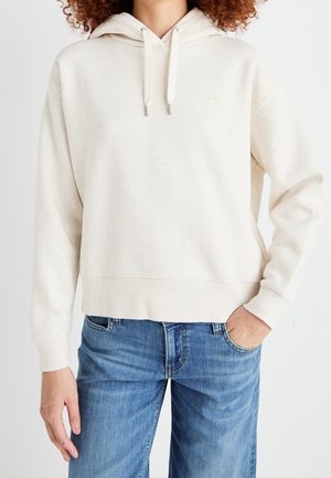 Personne portant un sweat à capuche beige clair avec cordons et un jean bleu en denim, une main dans la poche, sur fond blanc uni.