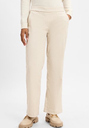 Beige, straight-leg trousers with a textured fabric and side pockets, paired with light-colored shoes featuring a chunky sole and gold accents.