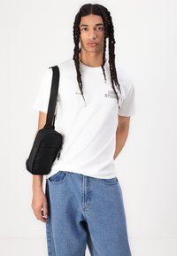 White t-shirt with "OBEY STUDIOS" text, paired with light blue denim jeans. A small black crossbody bag is worn over the shoulder.