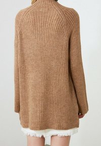 Maglione a coste color marrone chiaro con maniche lunghe indossato sopra una gonna bianca con orlo sfrangiato, visto da dietro.