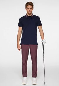 Mörkblå pikétröja med randig krage, vinröda smala byxor och vita sneakers. Personen håller i en golfklubba och bär en golfhandske.