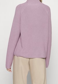 Suéter de punto lila con cuello alto, textura acanalada y hombros caídos, combinado con pantalones beige claros.