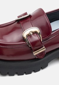 Burgundy patent leather loafer with a gold buckle detail, rubber tread sole, and contrasting black base. Smooth texture with stitched accents.