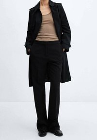 Trench-coat noir sur un débardeur beige, avec un pantalon noir sur mesure et des chaussures noires brillantes. La tenue présente une silhouette classique et des lignes épurées.