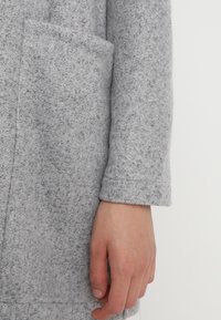 Rukáv šedého plstěného kabátu s viditelnou kapsou. Měkká textura, rovný střih a minimální šicí detaily. Ruka spočívající na tkanině.