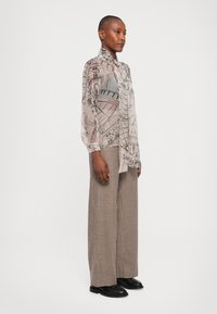 Durchsichtige, gemusterte Hemdbluse in Beige und Grau mit detaillierten architektonischen Drucken, kombiniert mit weit geschnittenen Hosen im Hahnentrittmuster.