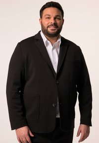 Blazer nero realizzato in tessuto liscio con revers a punta, due tasche frontali e chiusura a un bottone. Indossato sopra una camicia bianca.