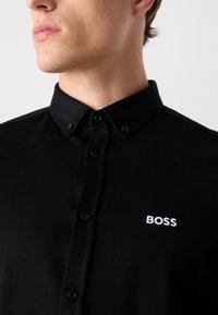 Black button-up shirt made of smooth fabric, featuring a classic collar, front placket, and white embroidered logo on the left chest.