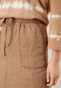 Persona che indossa una gonna di lino marrone con tasche frontali e vita con cordino, abbinata a un top a maniche lunghe marrone e bianco a tie-dye.