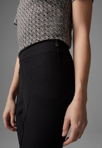 Des pantalons noirs ajustés présentent une texture lisse avec des poches arrière et une fermeture éclair sur le côté. Le haut à motifs met en avant un design géométrique dans des tons neutres.