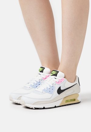 Baskets blanches Nike avec un swoosh noir, des accents bleu pastel et rose, portées pieds nus, vues de profil sur fond blanc.