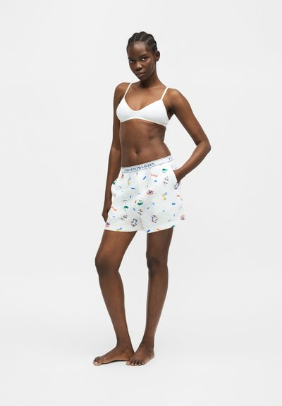 Γυναίκα που φοράει λευκό μπραλέ και λευκά σορτς με πολύχρωμα μικρά σχέδια και ζώνη μέσης με το λογότυπο Polo Ralph Lauren, όρθια ξυπόλητη σε ένα μονόχρωμο φόντο.