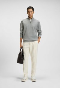 Felpa grigia con zip, pantaloni beige su misura e sneaker grigie e bianche. Tiene una borsa scura con manico superiore. Design semplice e moderno.