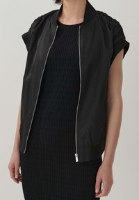 Svart bomberjacka med korta ärmar, tillverkad av glänsande tyg, med dragkedjestängning och rynkade axeldetaljer.