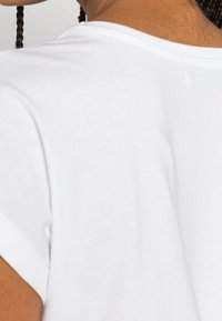 Biała bawełniana koszulka z prostym wzorem, z okrągłym dekoltem i krótkimi rękawami. Widok z tyłu pokazuje lekką fakturę i szczegóły szwów.