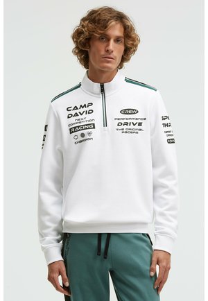 Young man with curly hair wearing a white zip-up sweatshirt with black racing-themed text and green striped athletic pants.