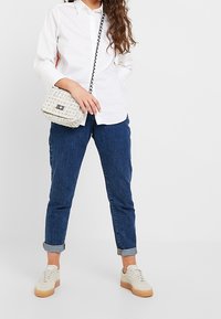 Camicia bianca a maniche lunghe con colletto, jeans blu con risvolto, sneakers beige e una piccola borsa a tracolla intrecciata con tracolla a catena.