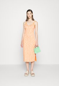 Vestido laranja com padrão, com alças finas e fenda lateral, combinado com uma mala verde clara e sandálias de plataforma beges. Textura suave.