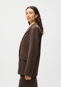 Brun skreddersydd blazer med en enkelt brystlomme og to frontlommer. Glatt tekstur, klippet revers og knappede mansjetter.