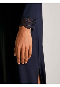 Abito in tessuto blu marina con maniche che presentano un polsino in pizzo ondulato. La mano mostra una pelle liscia con una semplice nail art, che mette in risalto i dettagli in pizzo.