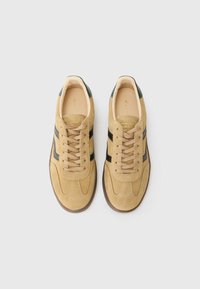 Tan suede sneakers with black stripes, round toe, beige laces, and green accents at the heel. Flat rubber outsole.