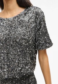 Koszulka z krótkim rękawem pokryta czarnymi, srebrnymi i szarymi cekinami. Miękka tekstura, luźny krój i szeroki dekolt podkreślają migoczące detale.