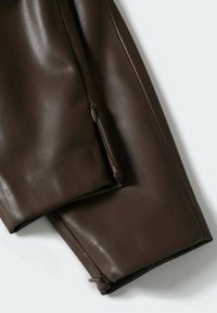 Bas de pantalon en cuir synthétique marron foncé montrant un détail de fermeture éclair sur la couture de la jambe, sur un fond clair uni.