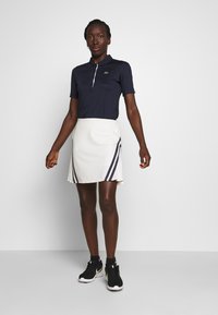 Polo shirt blu navy con colletto, gonna bianca con strisce blu navy e scarpe da uomo nere. Tessuto morbido e design aderente.