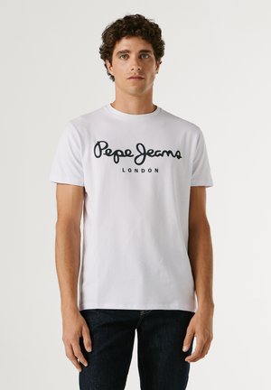 Mladi moški z kodrastimi lasmi, oblečen v belo majico z napisom "Pepe Jeans London" in temne kavbojke, stoji pred enostavnim svetlim ozadjem.