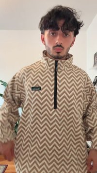 Suéter de media cremallera en beige con un patrón de zigzag blanco. Presenta una cremallera negra y un parche con el logo en el área del pecho izquierdo. Tejido texturizado.