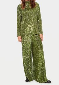 Donna che indossa un top verde a maniche lunghe con paillettes e pantaloni a gamba larga, in piedi davanti a uno sfondo semplice con scarpe nere visibili.