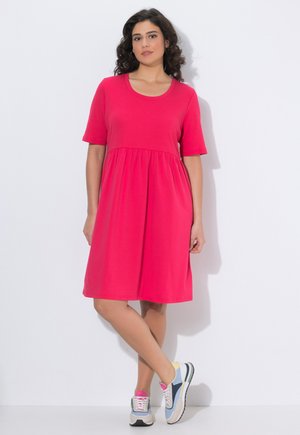 Femme aux cheveux bouclés et foncés, debout contre un mur blanc, portant une robe rose vif à manches courtes et longueur genou ainsi que des baskets multicolores.