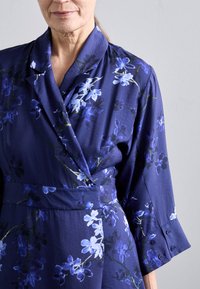 Tumši zilas krāsas apvērta kleita no gludas auduma ar ziedu rakstiem zilās un baltās toņos, ar šāla tipa apkakli un pogām uz piedurknēm.