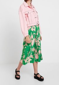 Rosa, kürz geschnittene Jeansjacke mit Knopfverschluss, kombiniert mit einem grünen Midirock mit Blumenmuster. Schwarze Plateau-Sandalen runden das Outfit ab.