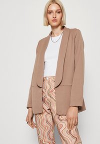 Beige sako s volným střihem, klopami a dlouhými rukávy, spárované s kalhotami s multikolórovým vlnitým vzorem a bílým žebrovaným topem.