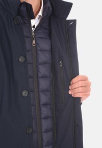 Giacca trapuntata blu navy con chiusura frontale a bottoni e cerniera, presenta uno strato interno rimovibile, tasche laterali e una texture liscia.
