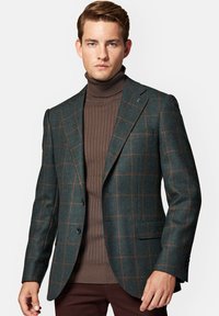 Ciemnozielony wełniany blazer w brązową kratę, z wykładanymi klapami, dwoma guzikami i kieszeniami, nałożony na brązowy sweter z golfem w prążki.